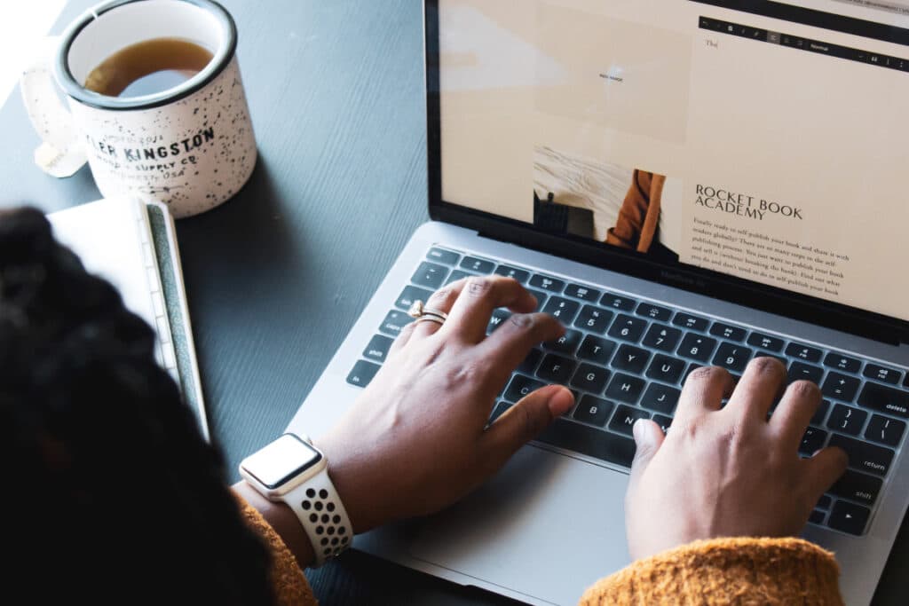over-the-shoulder photo of a woman typing on a macbook pro with a cup of tea sitting next to it